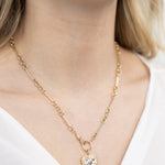 A person wearing a 14K gold pendant charm with a heart design, encrusted with abalone and a central sapphire, displayed against a white shirt neckline.