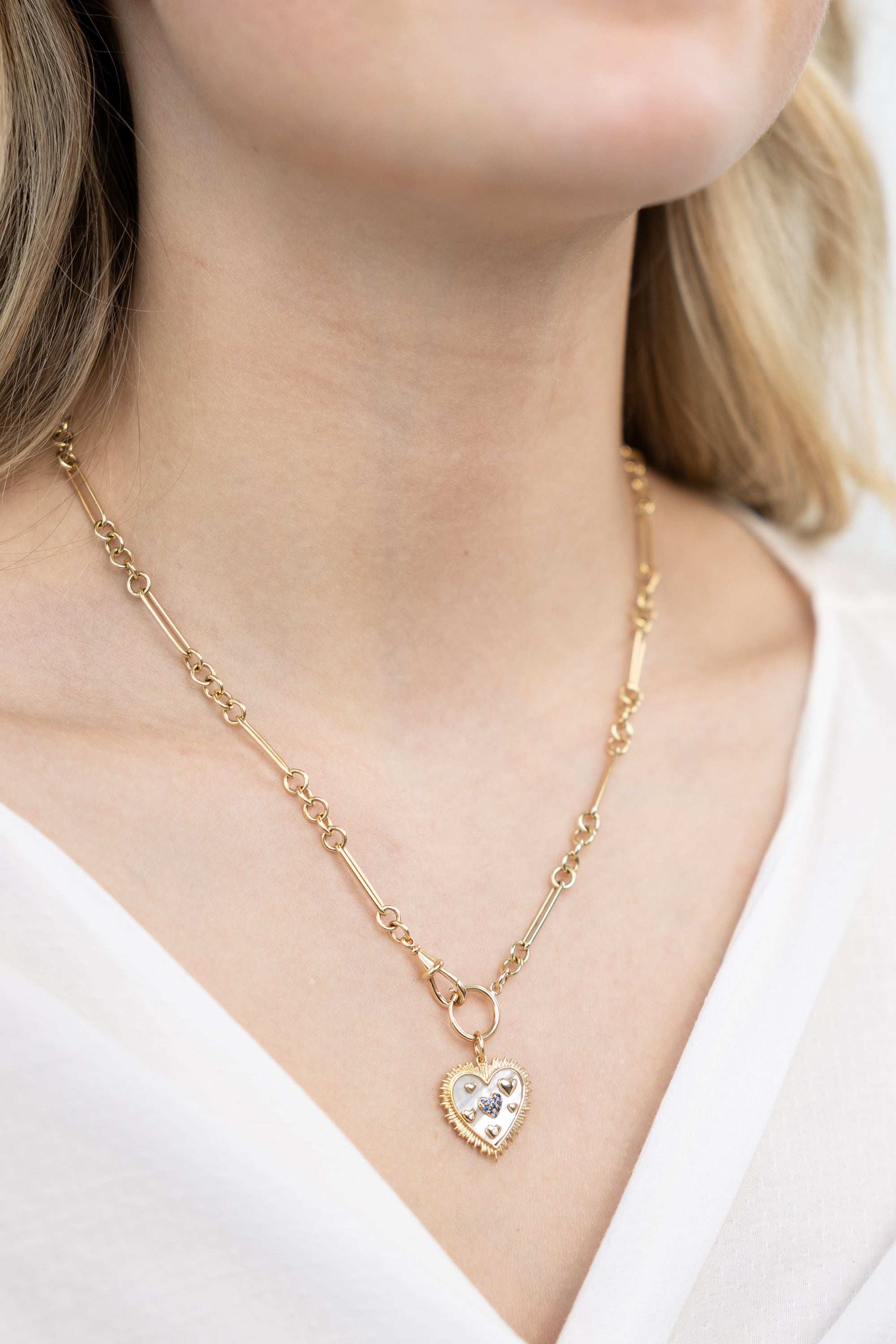 A person wearing a 14K gold pendant charm with a heart design, encrusted with abalone and a central sapphire, displayed against a white shirt neckline.