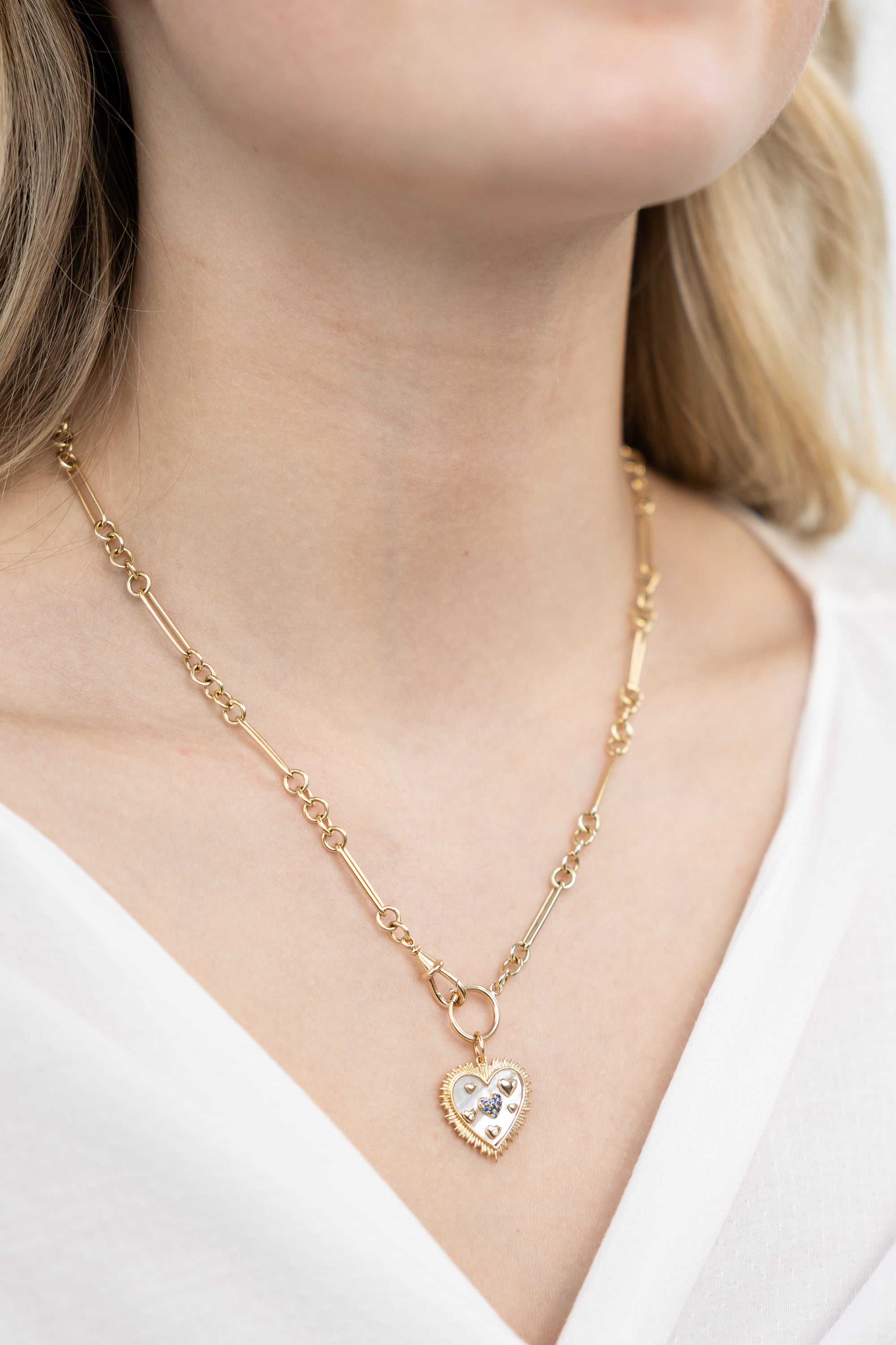 A person wearing a 14K gold pendant charm with a heart design, encrusted with abalone and a central sapphire, displayed against a white shirt neckline.