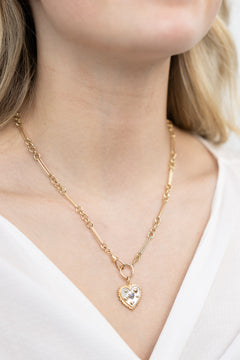 A person wearing a 14K gold pendant charm with a heart design, encrusted with abalone and a central sapphire, displayed against a white shirt neckline.
