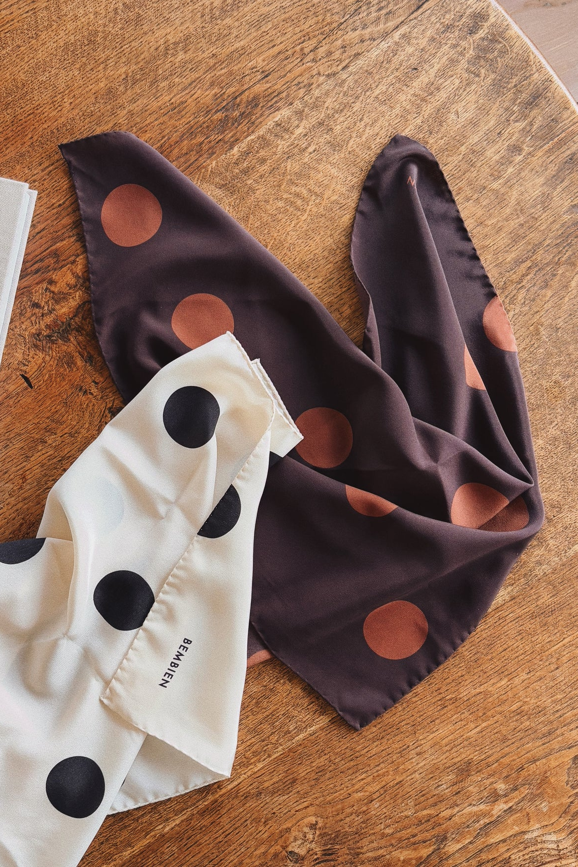 Two polka dot scarves, one white with black dots and the other brown with lighter brown dots, on a wooden surface.
