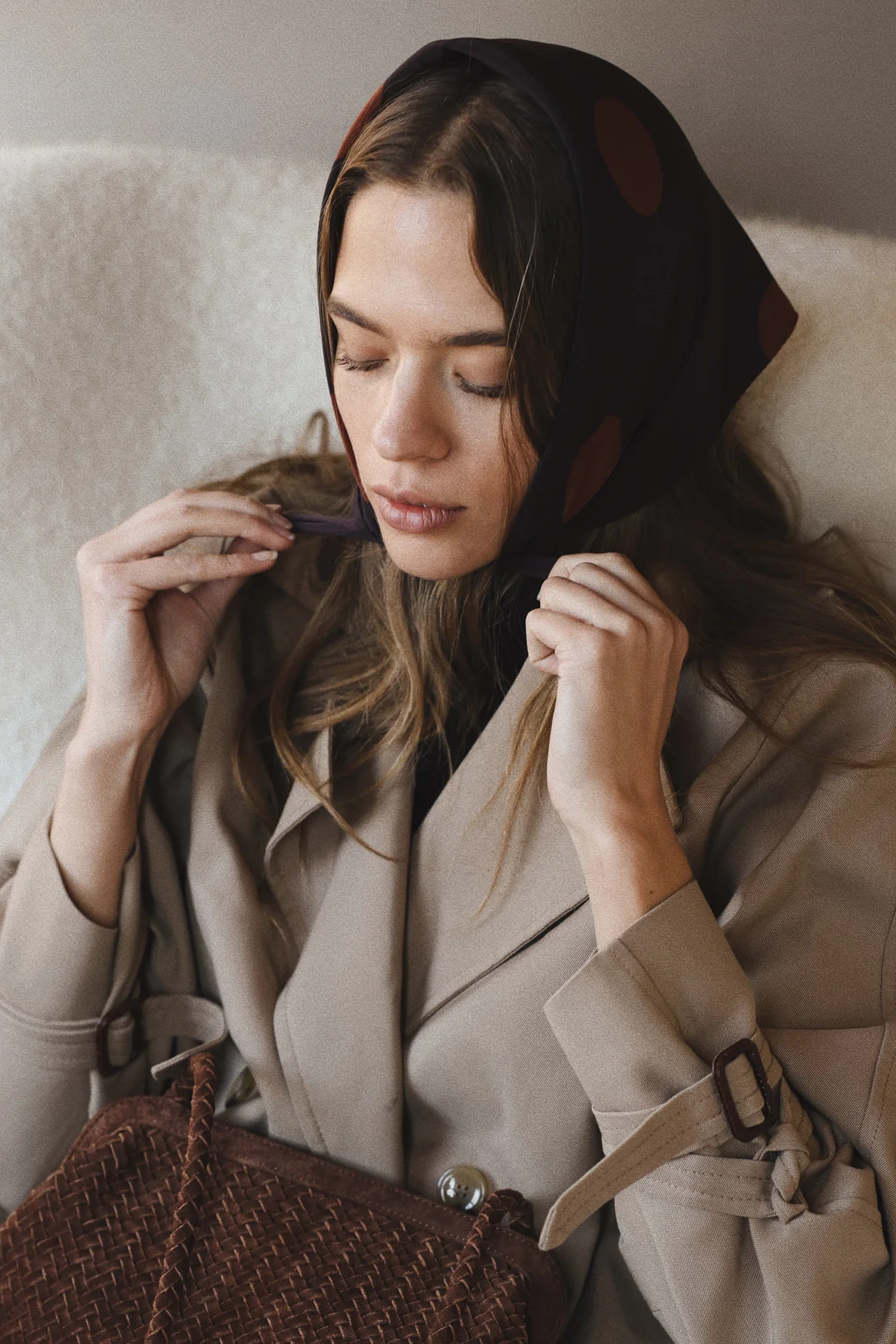 Woman in a beige coat sitting on a white couch, adjusting her scarf.