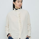Woman wearing a white blouse with ruffled details and dark pants against a plain background