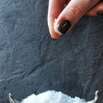 Hand sprinkling havsno salt flakes from a decorative salt cellar on a dark stone surface