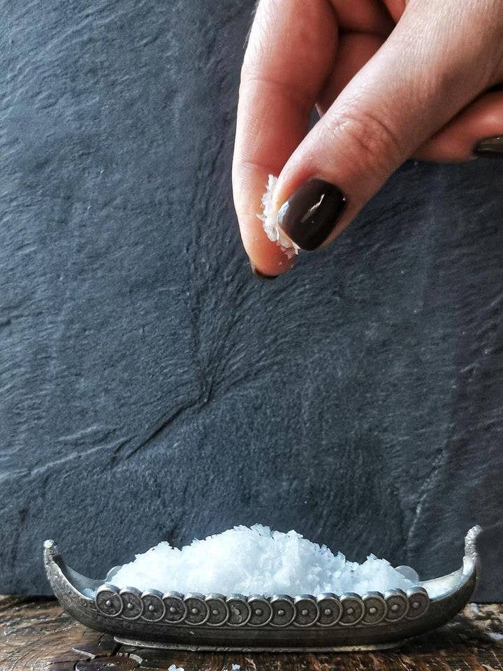 Hand sprinkling havsno salt flakes from a decorative salt cellar on a dark stone surface