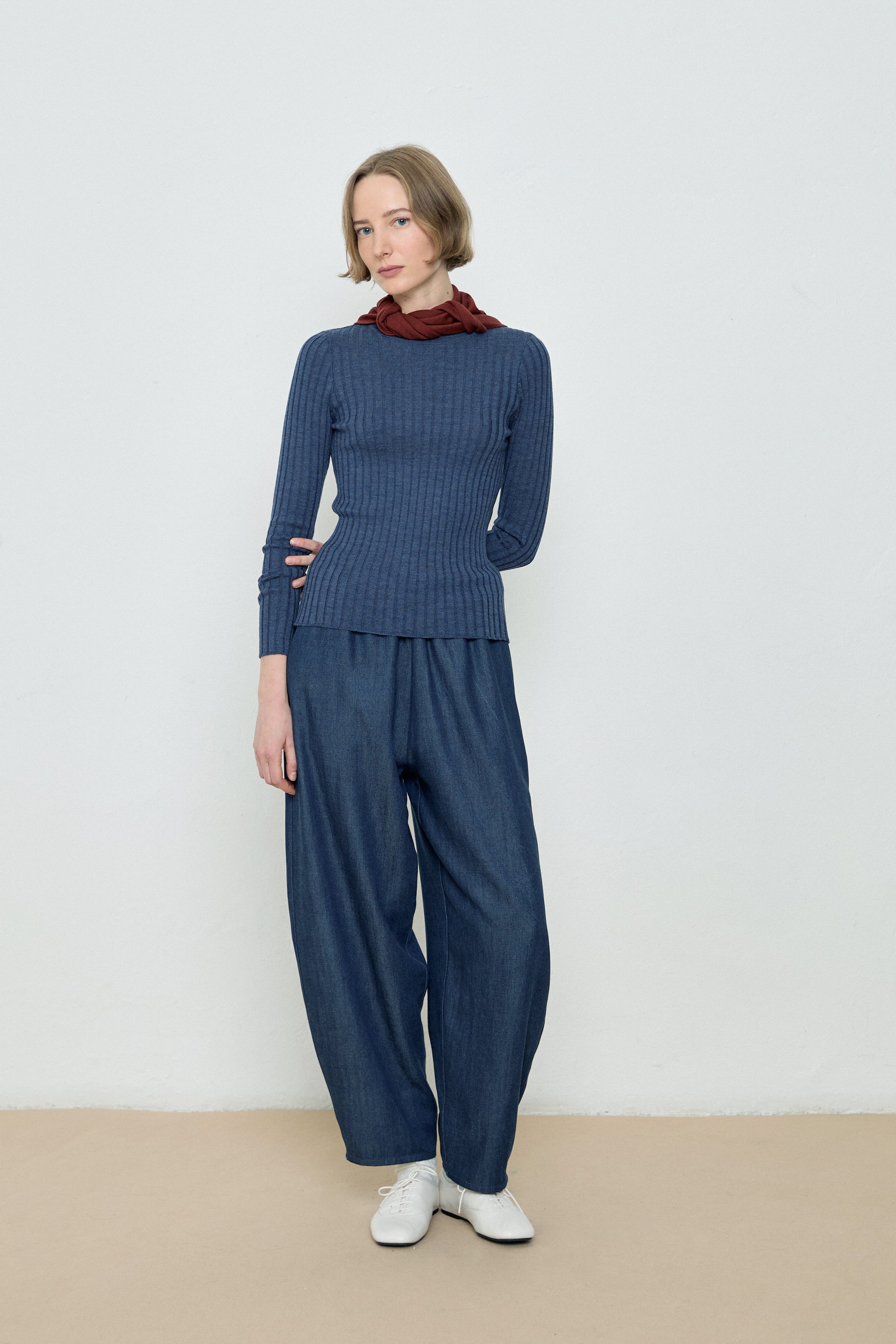 Woman wearing a blue ribbed sweater and matching pants with a red scarf against a white background