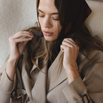 Woman in a beige coat sitting on a white couch, adjusting her scarf.