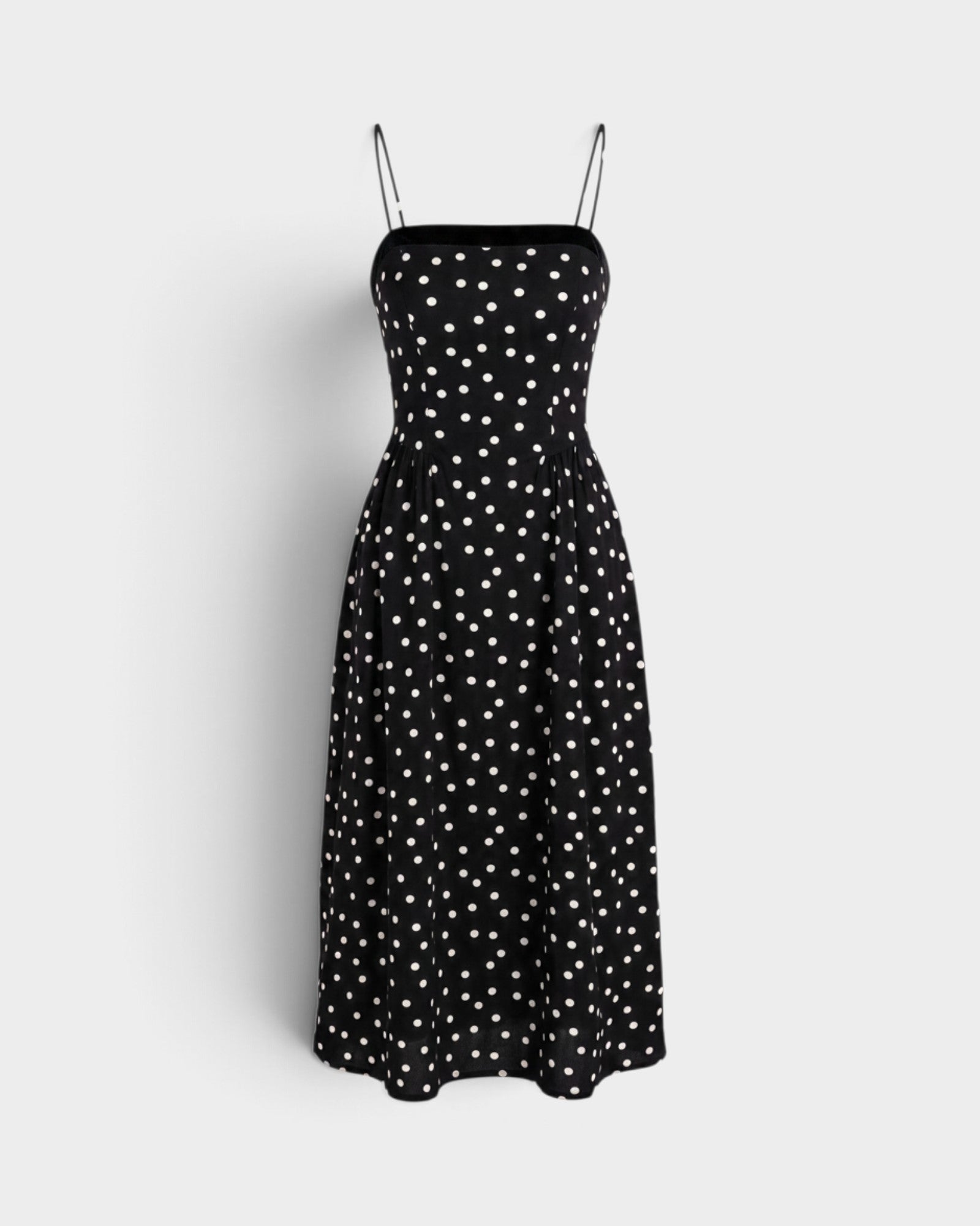 A black midi dress with a white polka dot pattern, featuring thin spaghetti straps and a fitted bodice, displayed against a light gray background.