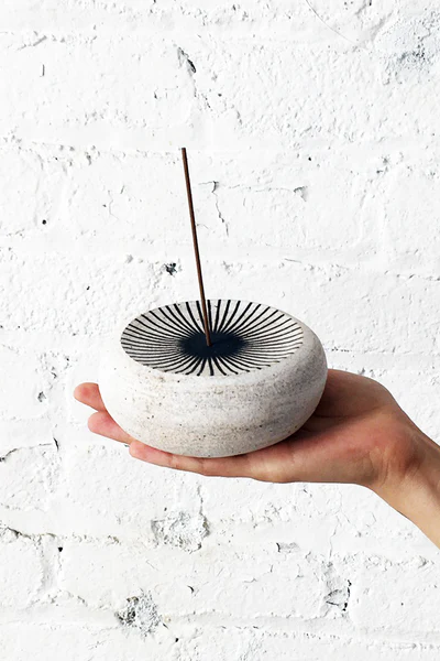 Hand holding a white incense burner with a black decorative element against a textured white background