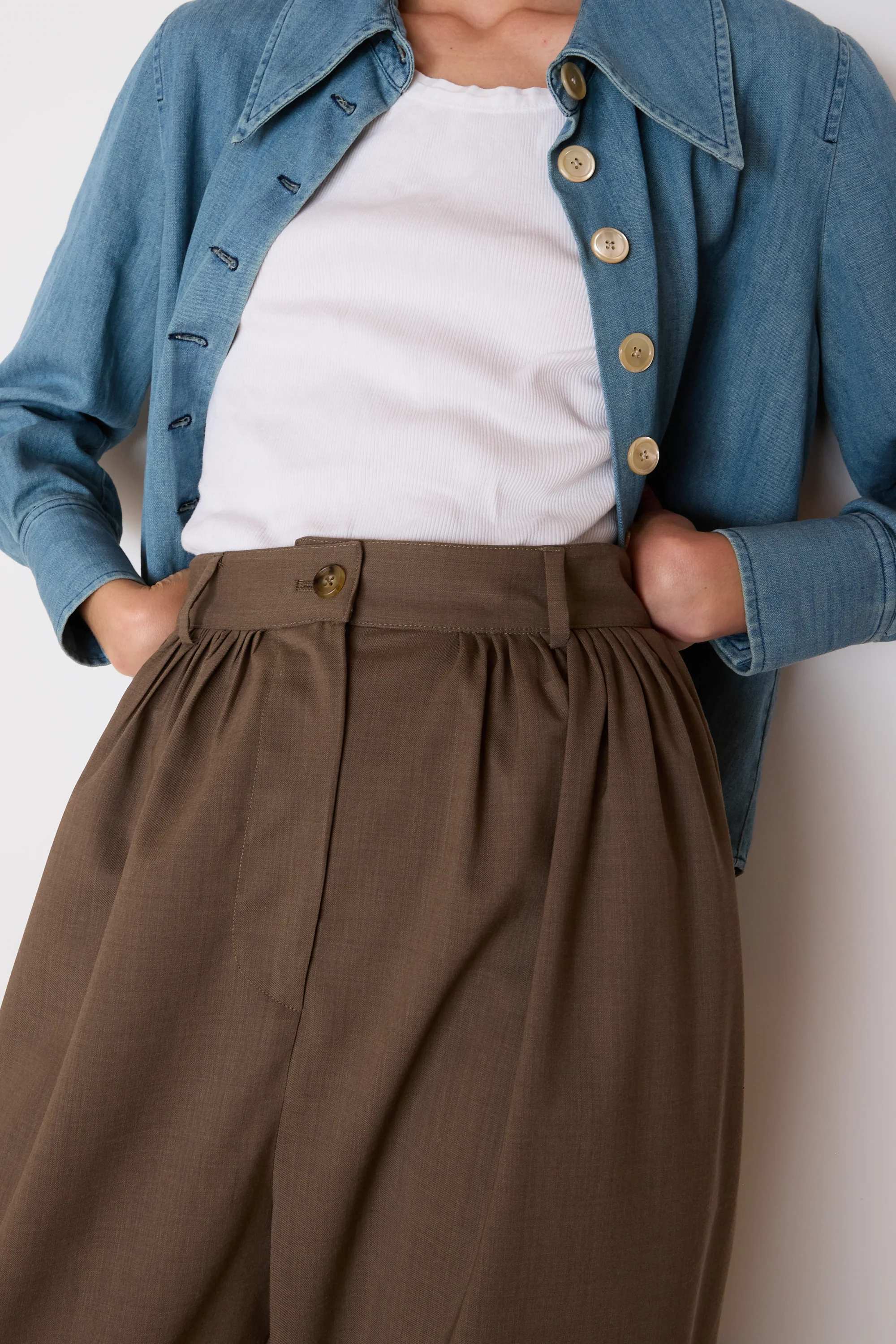 Person wearing a blue denim jacket, white shirt, and brown pants against a plain background