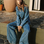 A woman wearing a light blue denim jumpsuit with wide-leg pants sits on stone steps outdoors. The background features a stucco wall, a large terracotta planter, and a window with curtains.