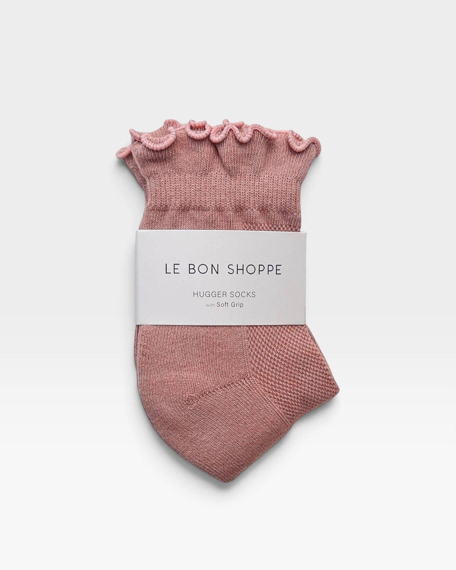 A pair of dusty pink knit huggie socks with a ruffled top edge, wrapped in a white paper band that reads 'LE BON SHOPPE HUGGER SOCKS with Soft Grip', on a light gray background.