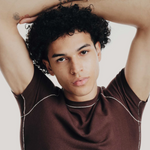 Man with tattoos on arms wearing a brown shirt against a plain background