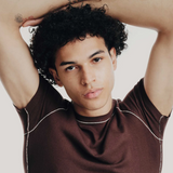 Man with tattoos on arms wearing a brown shirt against a plain background