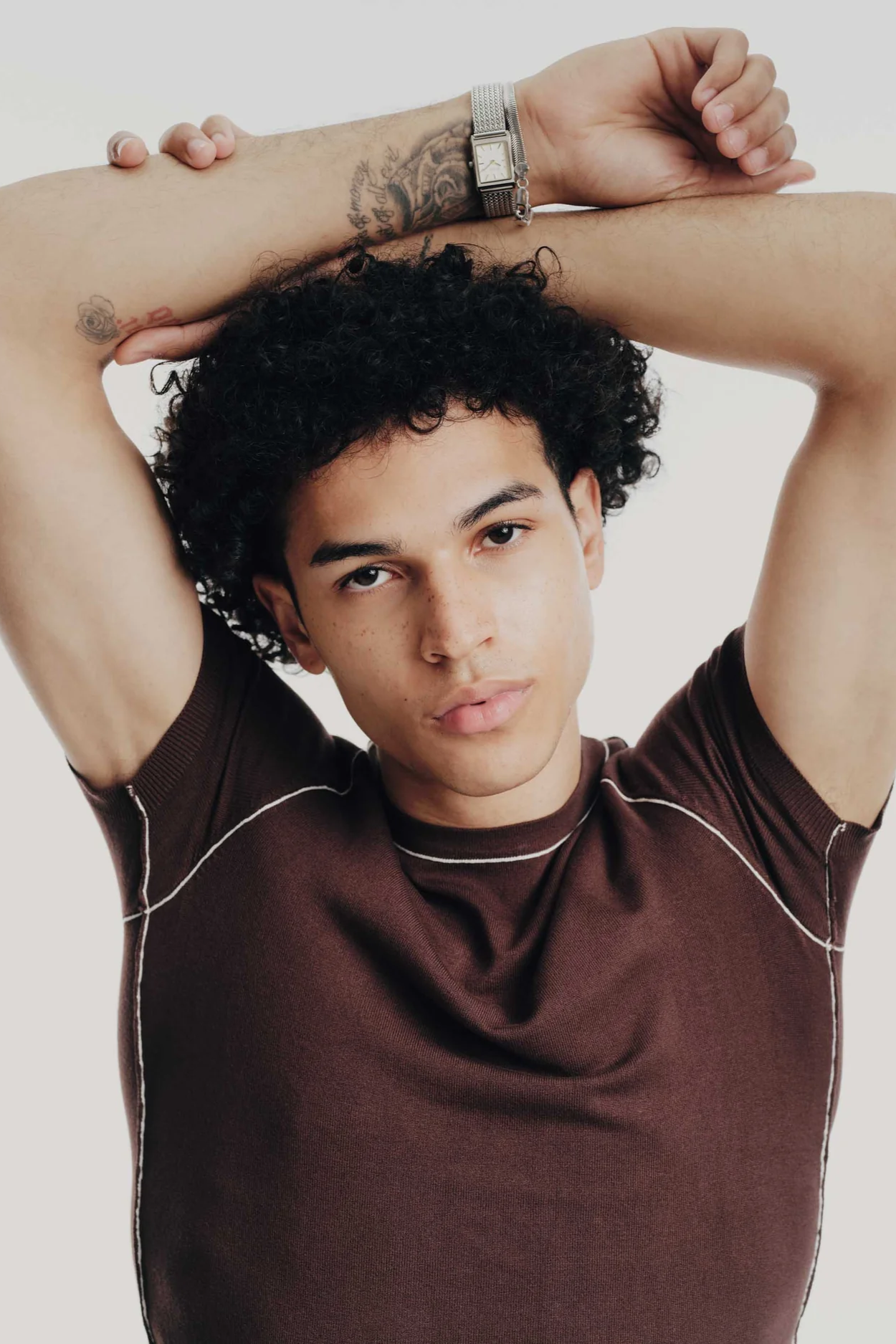 Man with tattoos on arms wearing a brown shirt against a plain background