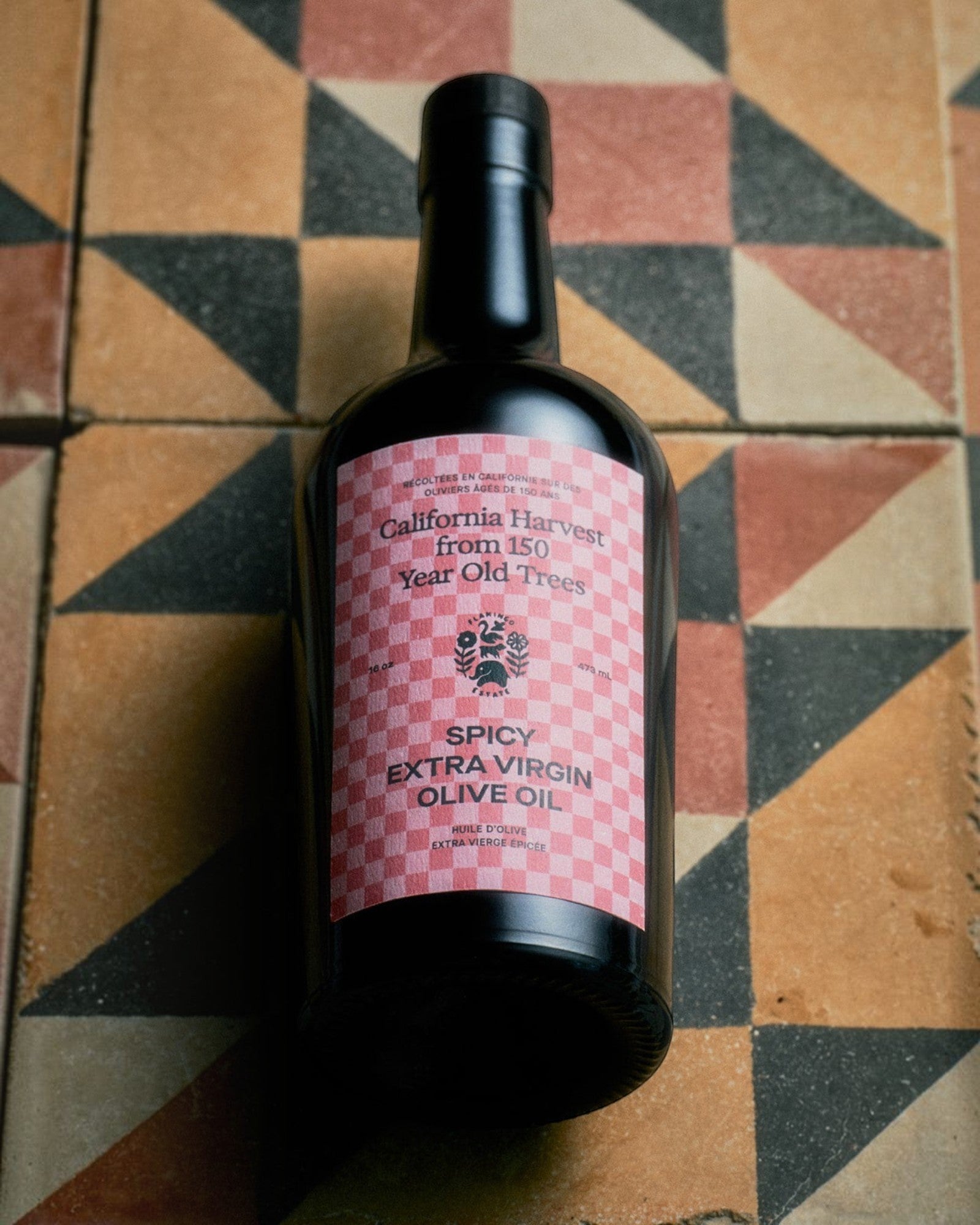 JONES + CO Spicy Extra Virgin Olive Oil in a dark glass bottle with a pink checkered label on a geometric tile floor.