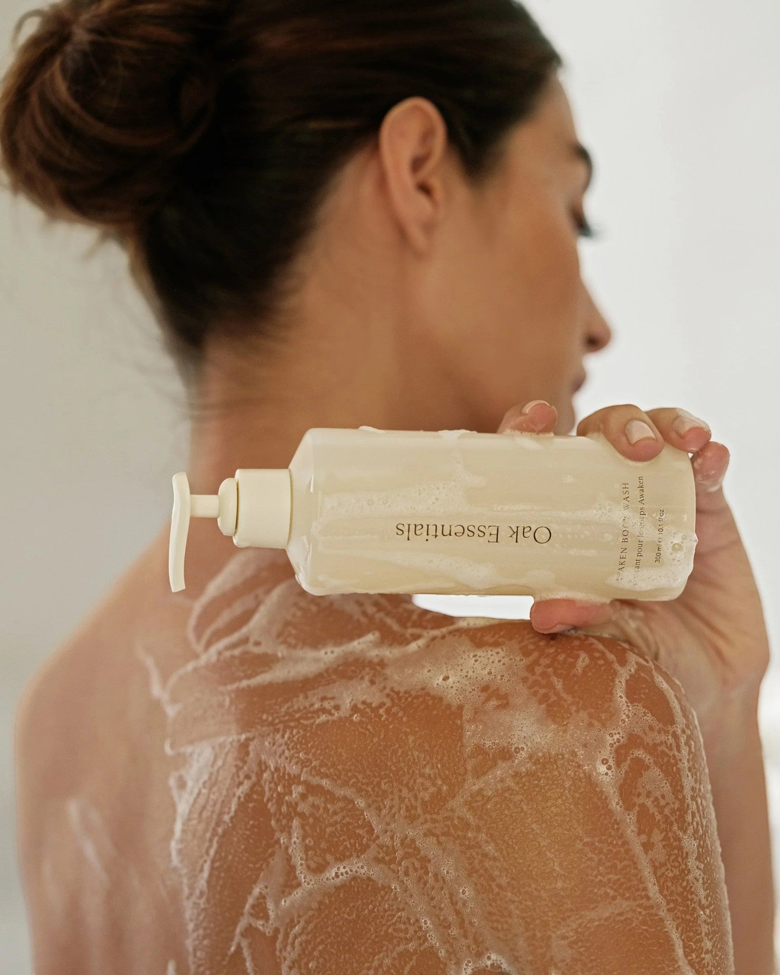 JONES + CO Awaken Body Wash bottle with botanical citrus scent, used by a woman in the shower.