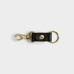 JONES + CO black leather keychain with a gold-tone metal clasp and ring on a light grey background.