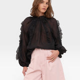 JONES + CO black sheer blouse with ruffled high neckline and voluminous sleeves, paired with pink wide-leg pants.