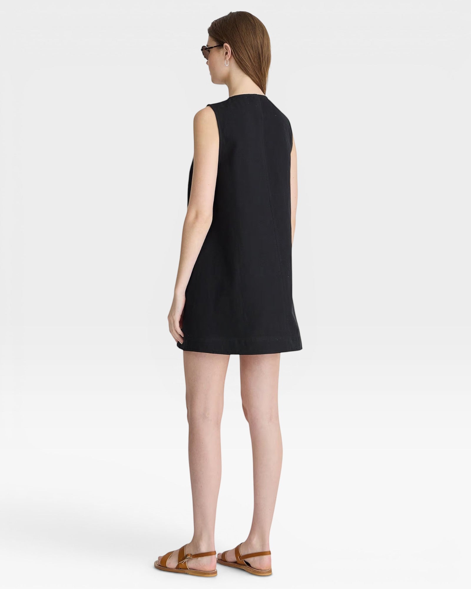 JONES + CO black sleeveless mini shift dress with a simple neckline and hem. Model wears sunglasses and sandals.