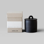 JONES + CO Pins & Needles petite candle in a black stoneware vessel with lid next to its beige cardboard box