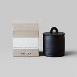 JONES + CO Pins & Needles petite candle in a black stoneware vessel with lid next to its beige cardboard box