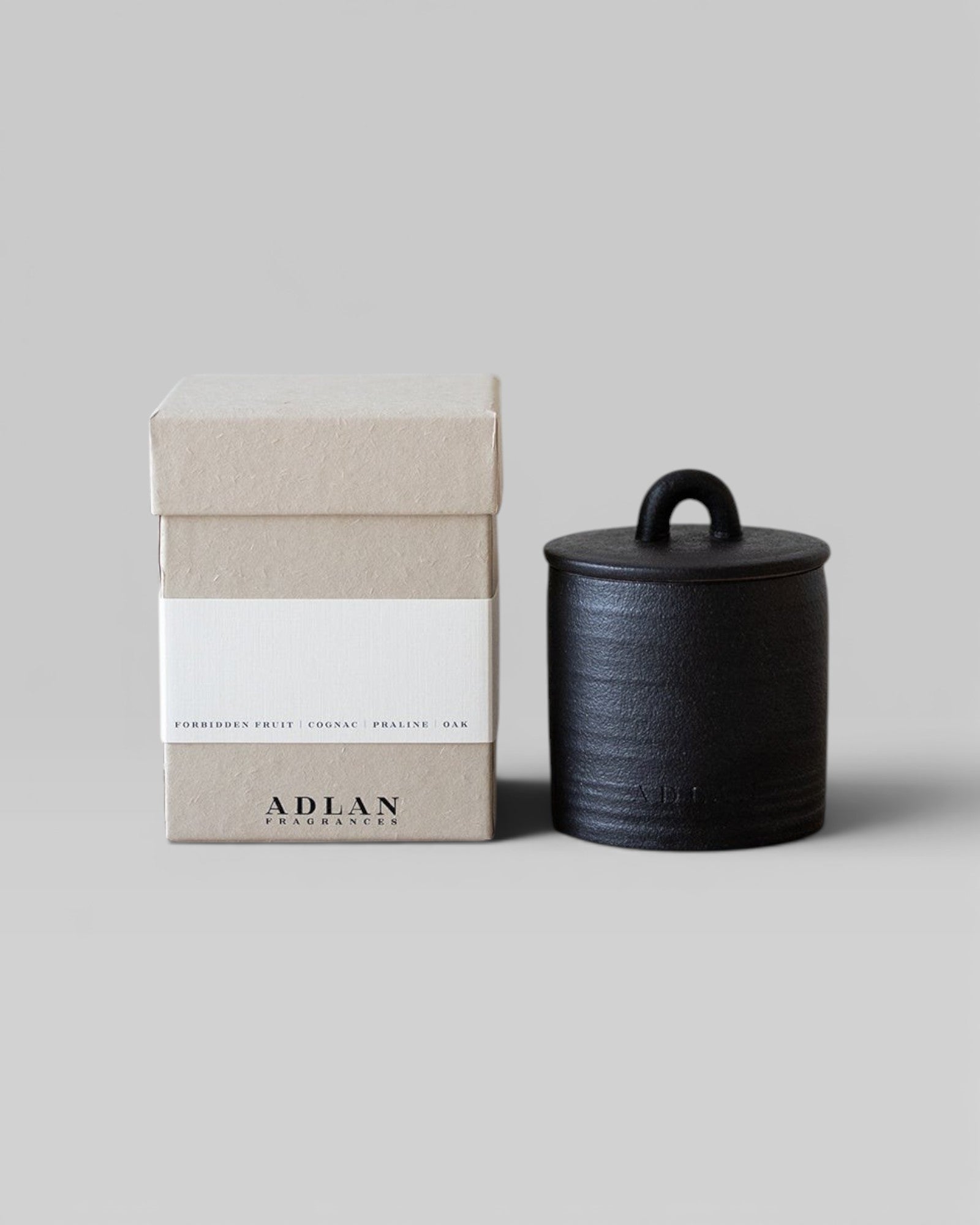 JONES + CO Pins & Needles petite candle in a black stoneware vessel with lid next to its beige cardboard box