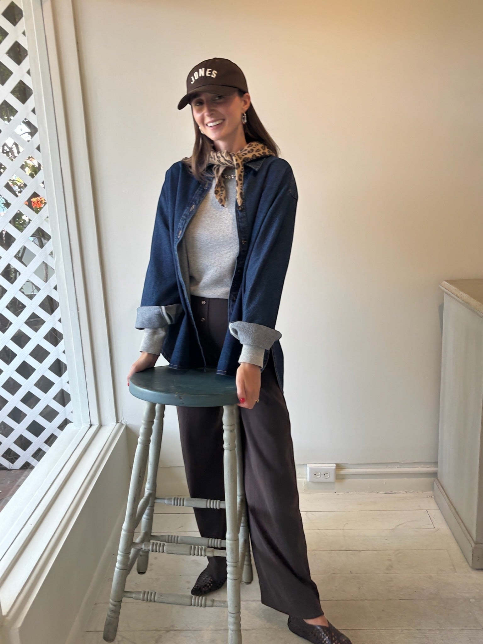 Woman wearing a brown JONES + CO baseball cap with white lettering, styled with a denim shirt and brown trousers.