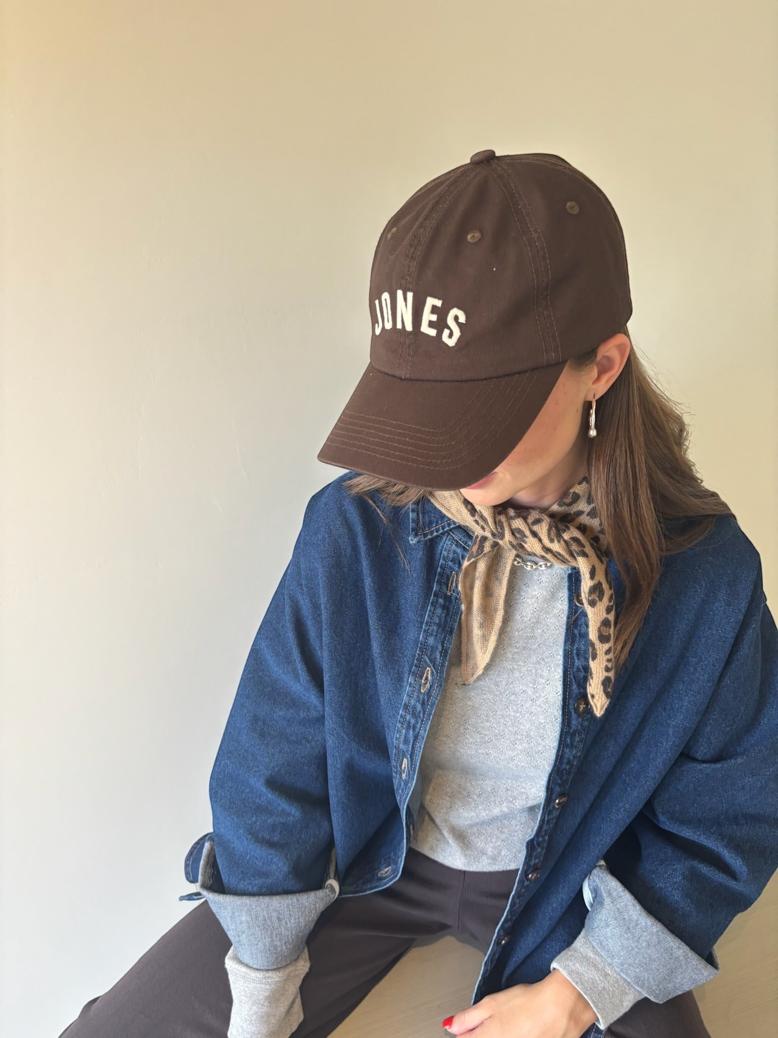 JONES + CO brown baseball cap with white embroidered lettering worn by a woman with a leopard print neck scarf