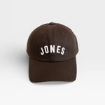 JONES + CO brown baseball cap with white embroidered JONES logo on the front, isolated on a white background.