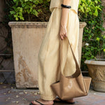 JONES + CO camel leather Tool Bag with exterior pockets and shoulder strap held by a woman in a pleated beige outfit.