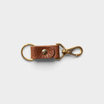 JONES + CO chocolate brown leather keychain with solid brass keyring and clip hardware on a light grey background.