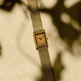 JONES + CO Virgil Small watch with a gold rectangular case, ochre dial, and woven mesh bracelet on a textured surface.