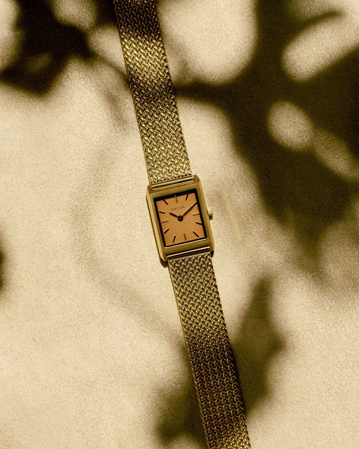 JONES + CO Virgil Small watch with a gold rectangular case, ochre dial, and woven mesh bracelet on a textured surface.