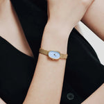 JONES + CO Jane Small watch with a gold oval case, sky blue dial, and woven mesh bracelet worn on a wrist.