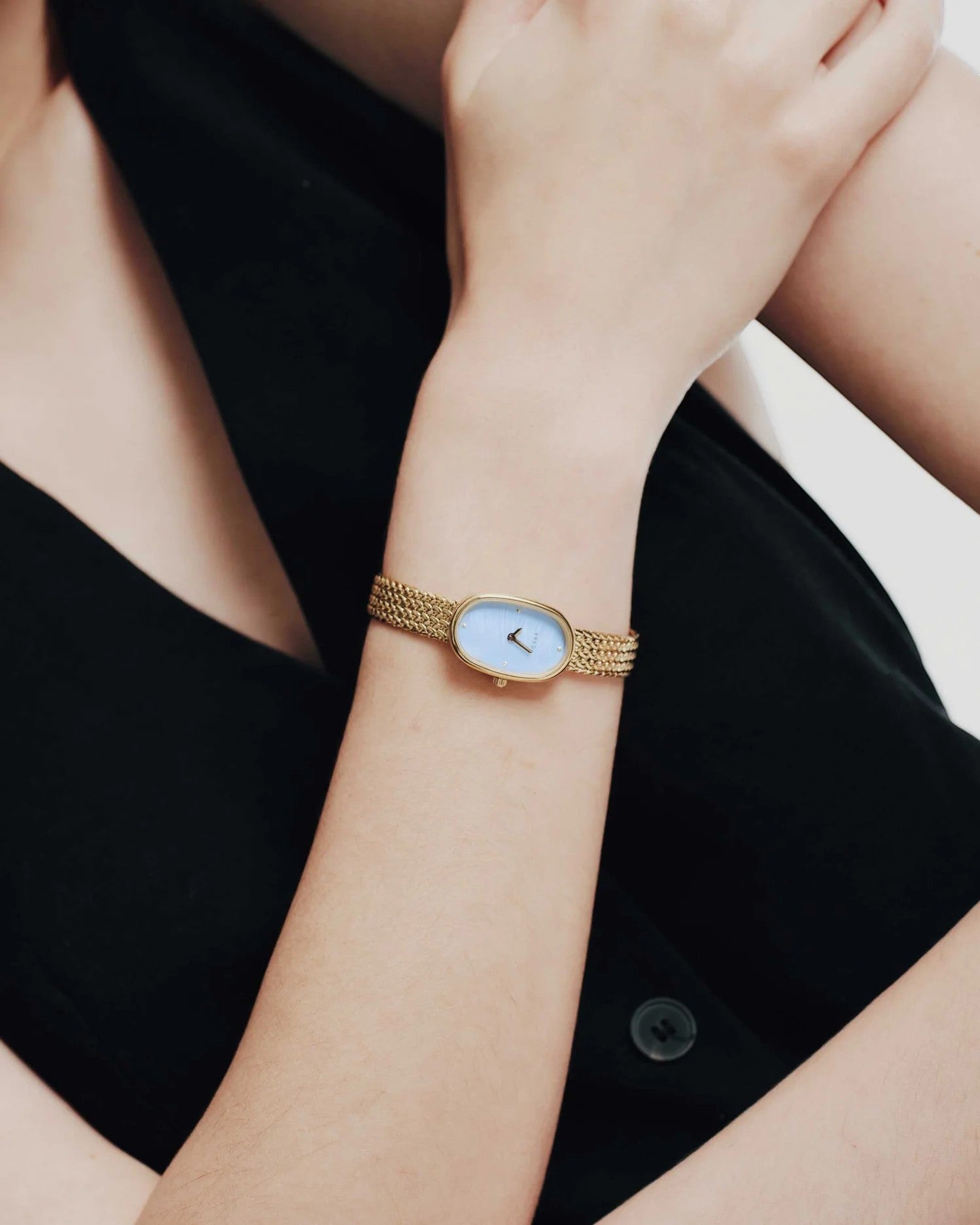 JONES + CO Jane Small watch with a gold oval case, sky blue dial, and woven mesh bracelet worn on a wrist.