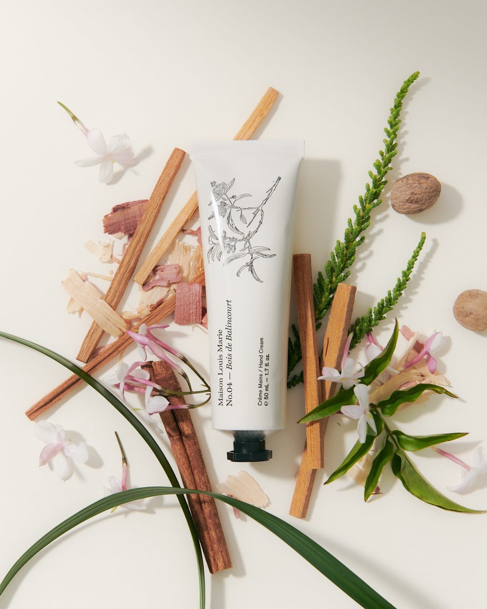 JONES + CO No.04 Bois de Balincourt hand cream tube with botanical illustrations, surrounded by wood chips, cinnamon sticks, and jasmine flowers.