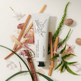 JONES + CO No.04 Bois de Balincourt hand cream tube with botanical illustrations, surrounded by wood chips, cinnamon sticks, and jasmine flowers.