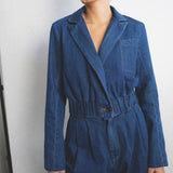 JONES + CO indigo denim wide-leg jumpsuit with lapel collar, elasticated waist, and front pleats. Model wears statement earrings.
