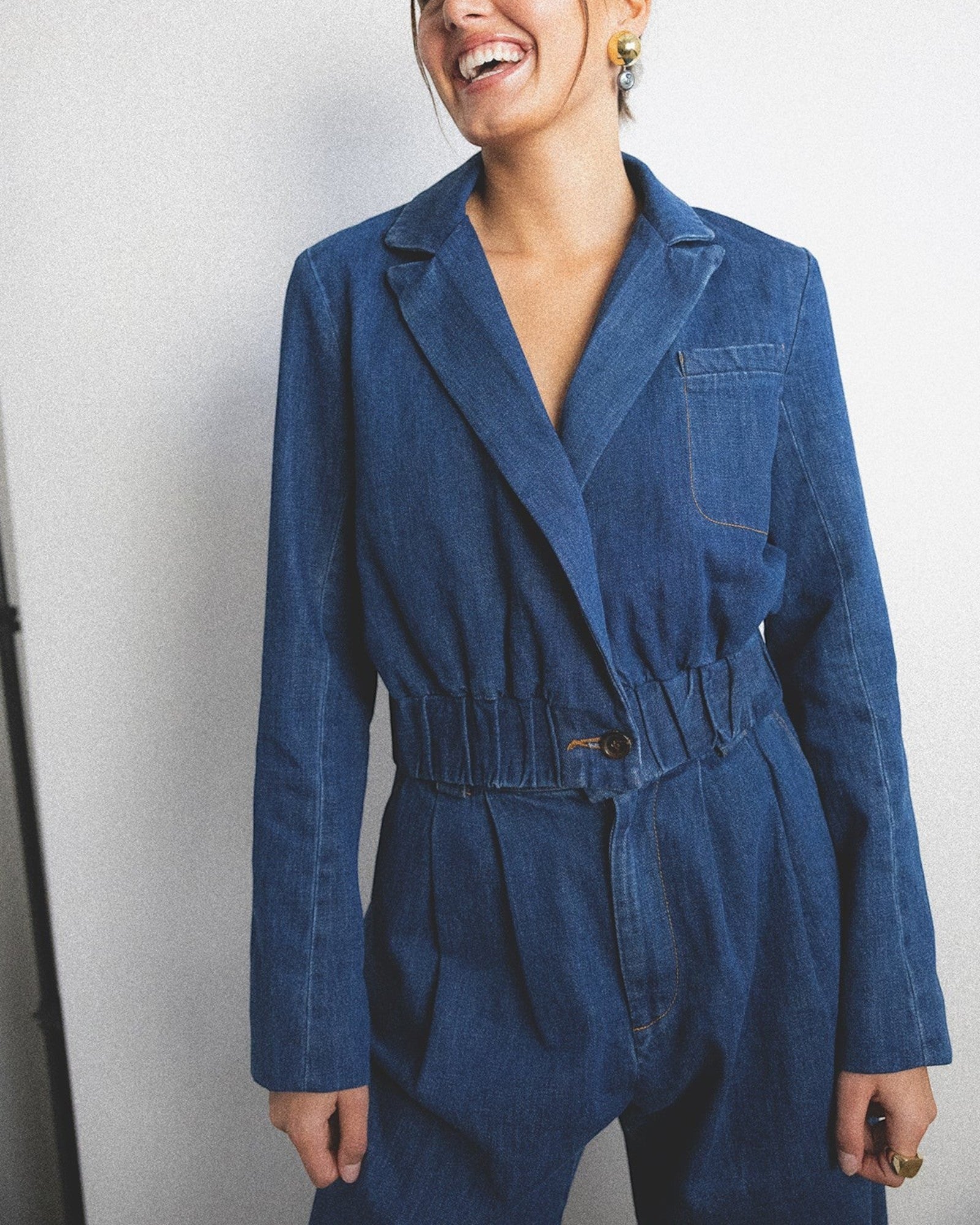 JONES + CO indigo denim wide-leg jumpsuit with lapel collar, elasticated waist, and front pleats. Model wears statement earrings.
