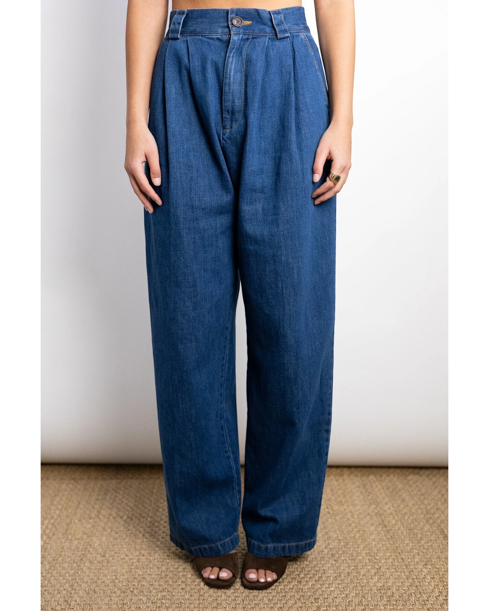 JONES + CO indigo denim Hede pants with high waist, wide leg, front pleats, and side pockets. Model wears them with a gold ring.