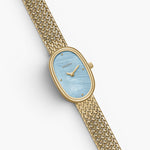 JONES + CO Jane Small watch with a gold oval case, sky blue mother of pearl dial, and woven gold mesh bracelet.