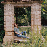 JONES + CO Jess Blouse in Muir Tartan with pintuck details, worn by a woman sitting in a stone garden archway.