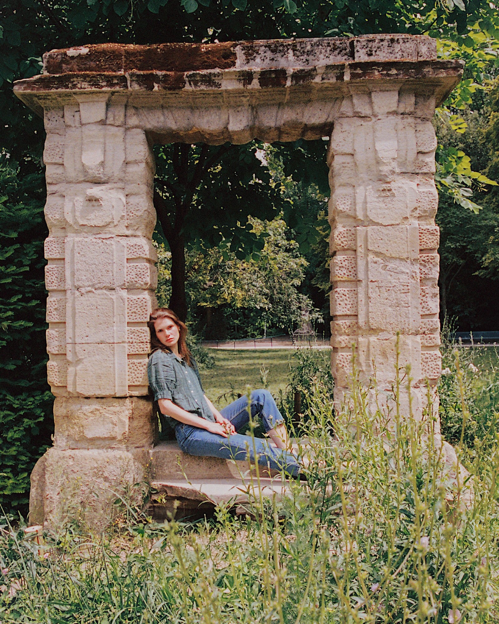 JONES + CO Jess Blouse in Muir Tartan with pintuck details, worn by a woman sitting in a stone garden archway.