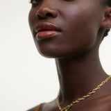 JONES + CO La Laque Tinted Lip Oil in a warm coral shade, worn by a model with dark skin and gold jewelry.