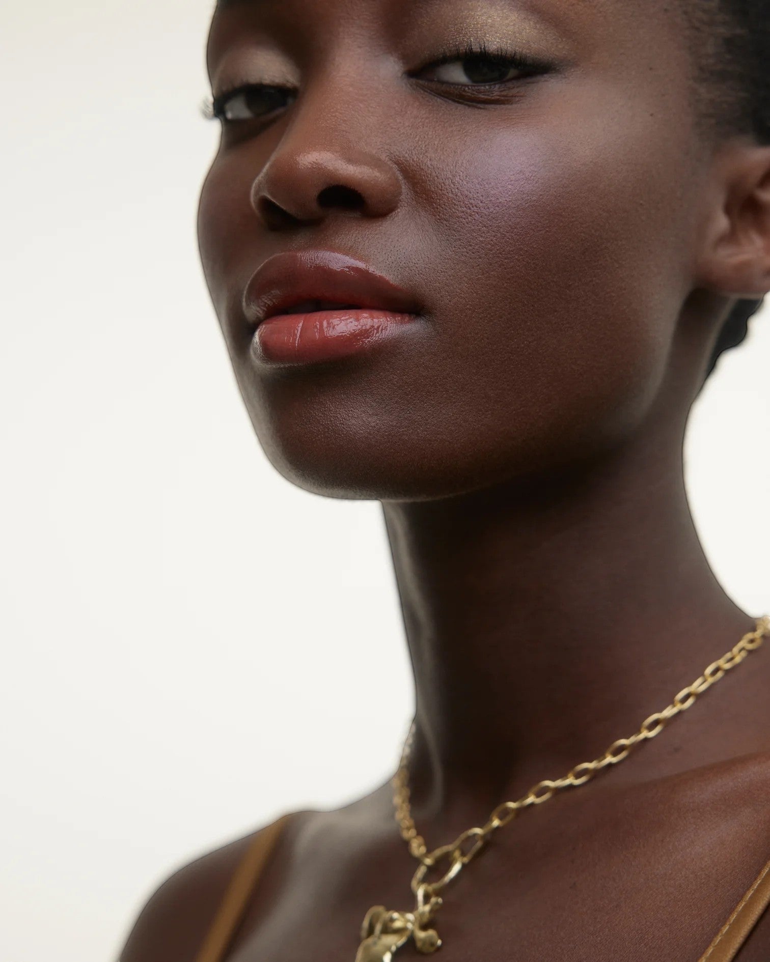 JONES + CO La Laque Tinted Lip Oil in a warm coral shade, worn by a model with dark skin and gold jewelry.