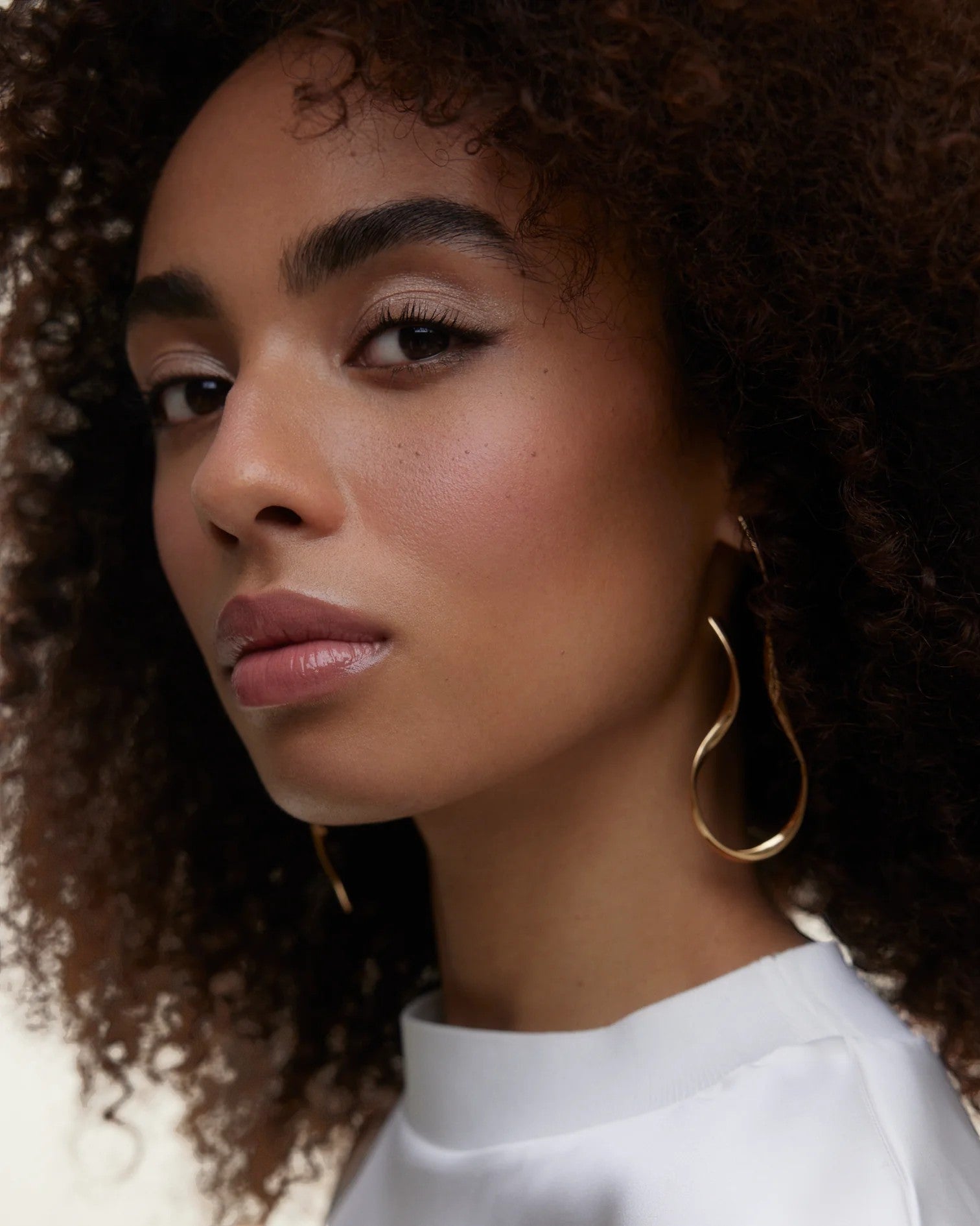 JONES + CO La Laque Tinted Lip Oil in a rosy nude shade, worn by a model with curly hair and gold earrings.