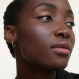 JONES + CO La Laque Tinted Lip Oil in a sheer, luminous rosy-nude shade, applied to a model with glowing skin and subtle shimmer.