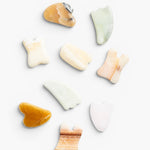 JONES + CO Natural Gua Sha Stones in various shapes and earthy tones like jade green, peach, and amber for depuffing and improving circulation.
