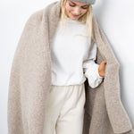 JONES + CO oatmeal alpaca wool blend throw blanket draped over a woman wearing a grey beanie and white sweater.